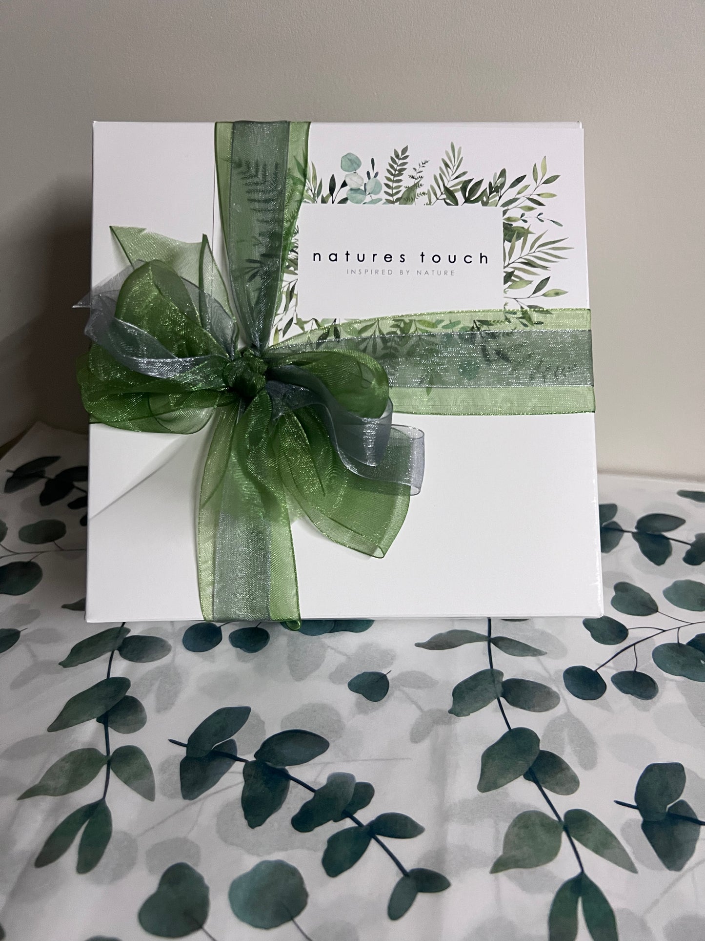 
                  
                    A white square gift box with botanical leaf illustrations and gift card, tied with a sheer green and silver ribbon and bow
                  
                